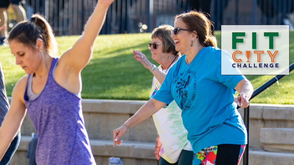 Three women exercising outdoors on a sunny day, smiling and moving energetically. In the corner, a logo reads "FIT CITY CHALLENGE" with a green and orange design. Grass and stairs are visible in the background.